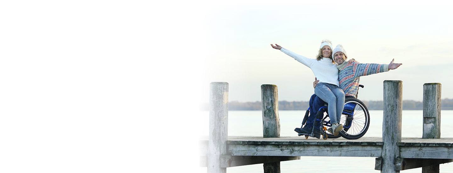 Man in wheelchair holds wife on lap on a dock.